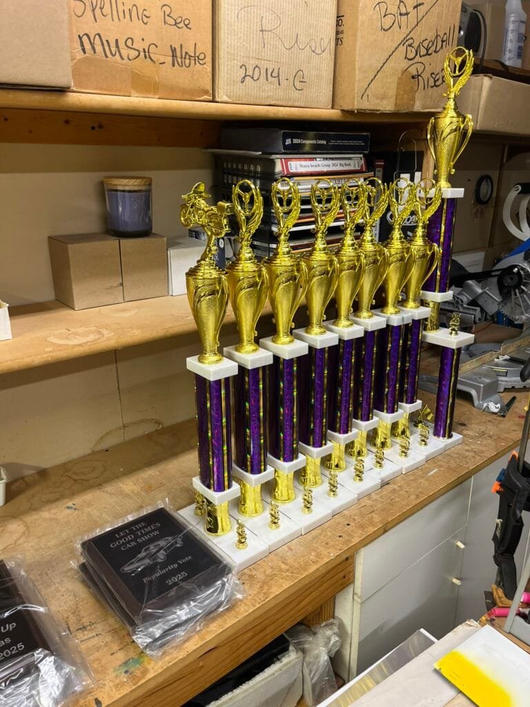 Row of gold trophies with purple stems lined up on a wooden workbench in a workshop, background boxes and tools visible.