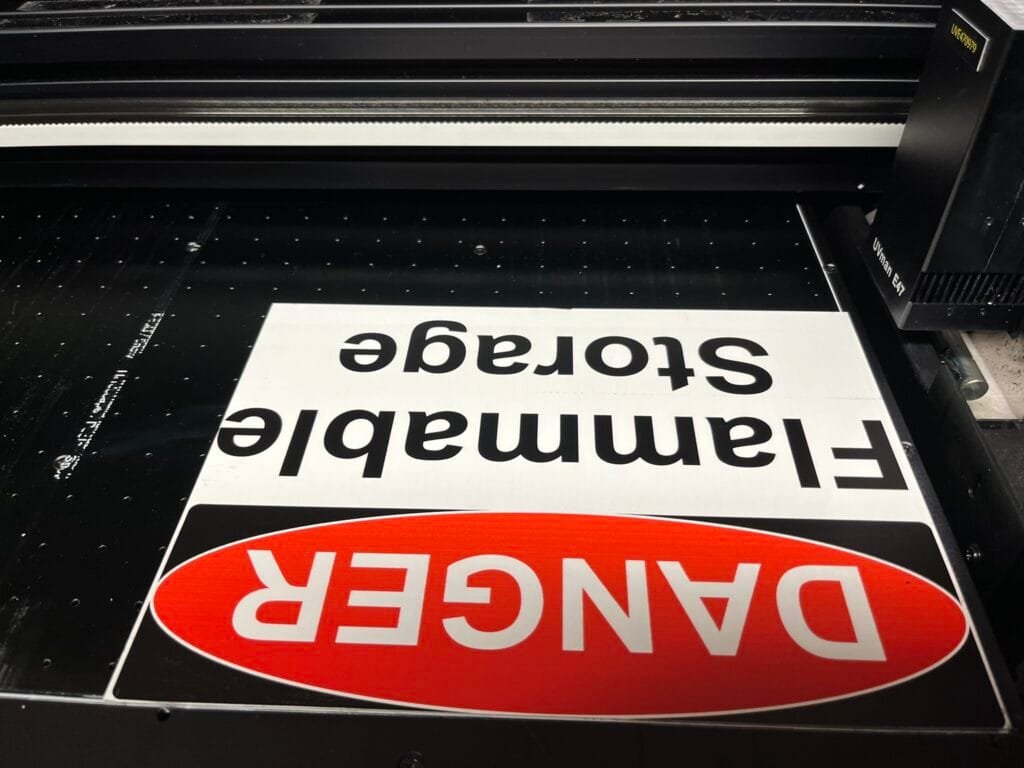 Upside-down sign with bold black text reading'Emergency' on a black machine bed, with a red warning graphic partially visible at the bottom.