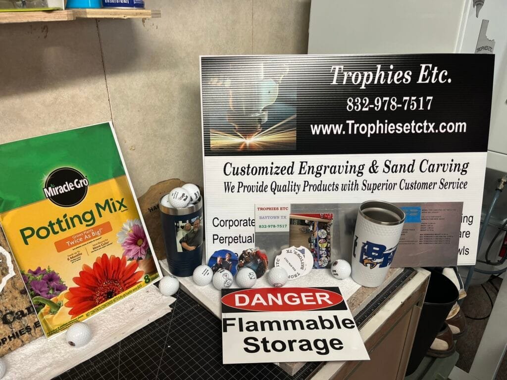 Cluttered workbench with a large Trophies Etc. sign, contact info, and engraving ads, plus scattered golf balls and mugs on the counter.