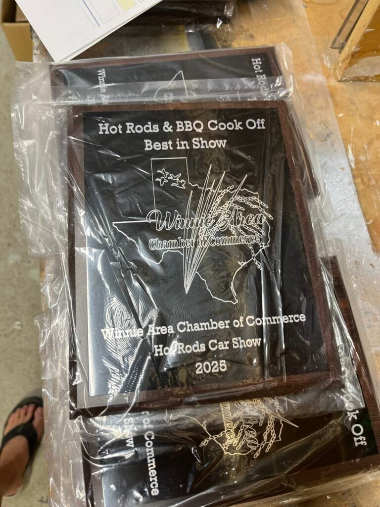 Plastic-wrapped award plaque on a table; reads'Hot Rods & BBQ Cook Off Best in Show' with 'Chamber of Commerce' at the bottom.