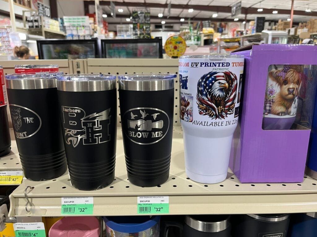 Row of insulated tumblers on a store shelf, including a black tumbler with 'BLOW ME' and a white patriotic design tumbler.