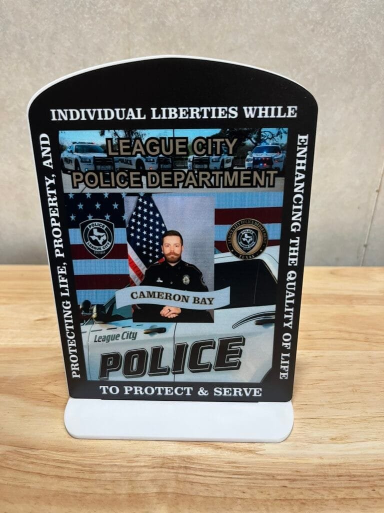 Informational sign for League City Police Department featuring a uniformed officer, American flag, and police cars in a banner display, with 'To Protect & Serve' text at the bottom.