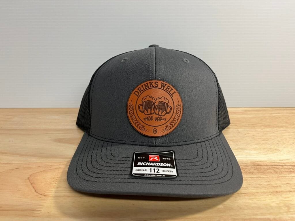 Gray trucker hat with a brown circular patch reading 'DRINKS WELL' on the front, placed on a light wood surface. Patch shows beer mugs motif, mesh back visible.