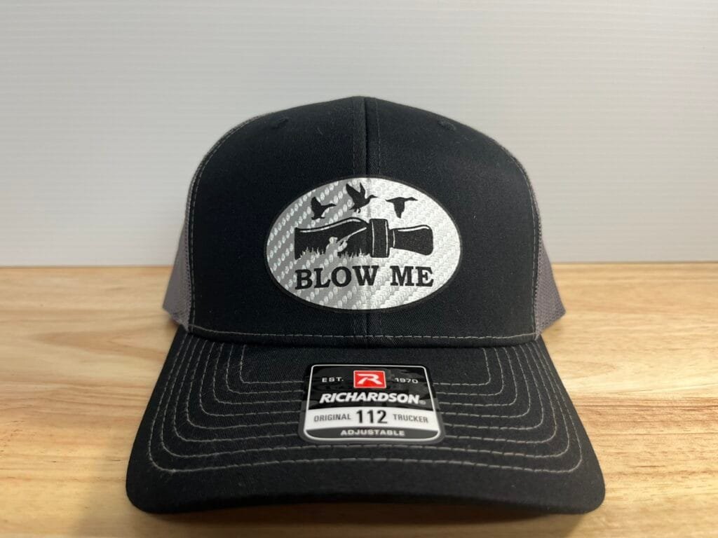 Black Richardson 112 adjustable trucker cap on a wooden surface, featuring a round patch with a flashlight illustration, birds, and the text 'BLOW ME' in black letters on a metallic background.",