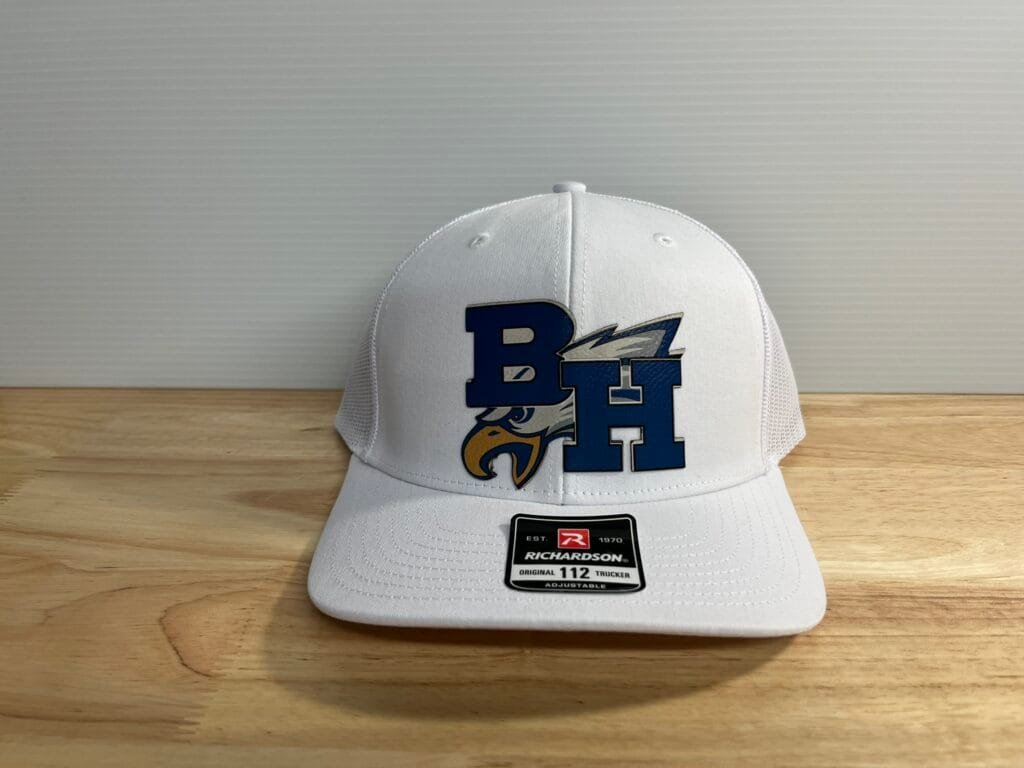 White trucker cap with a blue block-letter logo and eagle graphic on the front, resting on a wooden surface with an adjustable fit sticker on the brim.