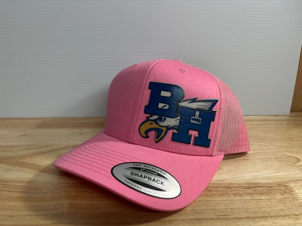 Pink trucker-style cap with a bold blue graphic on the front and a circular 'Snapback' sticker on the brim, resting on a wooden surface.