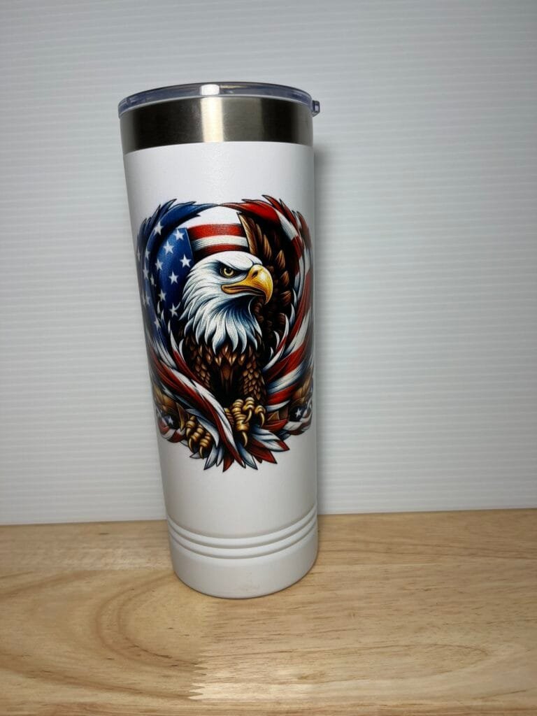 White stainless steel tumbler with a detailed eagle and American flag motif on a white background, sitting on a wooden table.