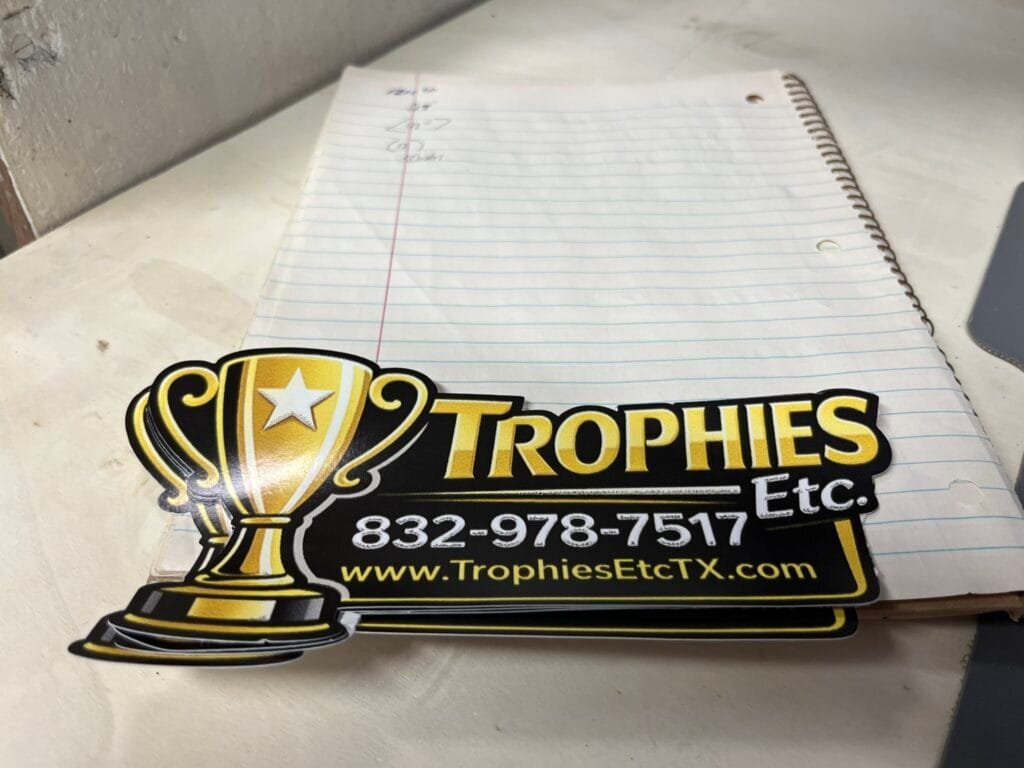 Promotional sticker on a notebook featuring a trophy logo and contact website for Trophies Etc.