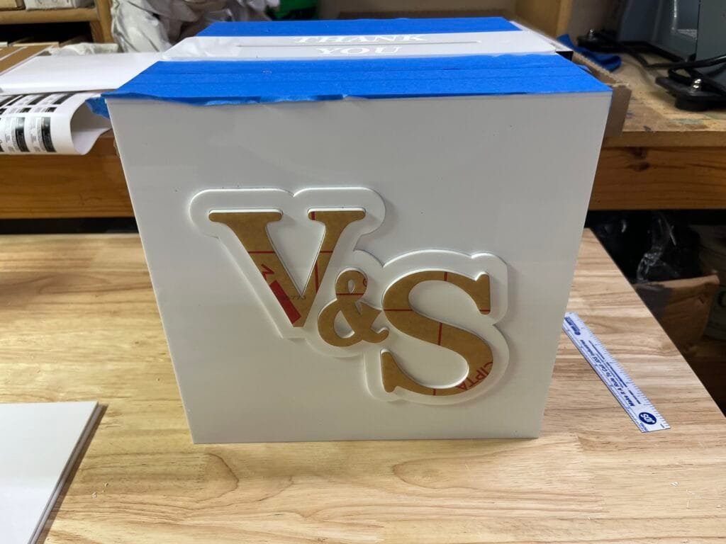 White gift box with blue painter's tape on top and a 'THANK YOU' card, on a workbench.